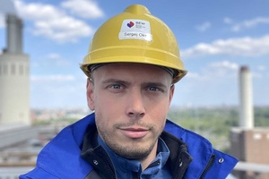 Selfie eines Mannes mit gelbem Bauhelm und blauer Jacke vor Industrieschornsteinen