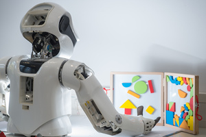 Rückansicht eines humanoiden Roboter-Oberkörpers, der auf einem Tisch steht und mit einer Hand nach einer Form auf einer Magnettafel greift