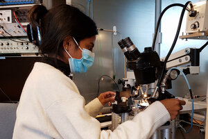 Frau mit Laborkittel und Mund-Nasen-Schutz blickt in ein Mikroskop