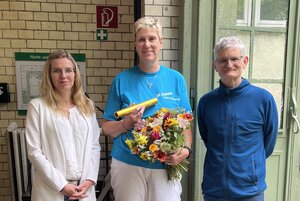 Drei Personen stehen nebeneinander und blicken zur Kamera, die mittlere hält einen großen Blumenstrauß