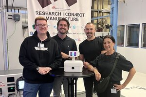 Vier Personen stehen um einen Stehtisch herum und schauen in die Kamera. Auf dem Tisch steht ein kleiner Roboter.