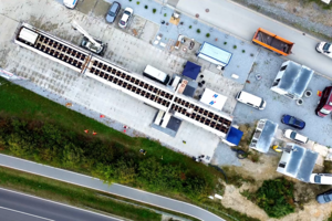 Luftaufnahme eines betonierten Areals neben einer Straße, auf dem sich eine Brücke sowie Transporter, Container und ein kleiner Baukran befinden