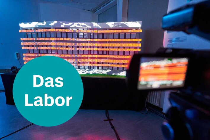Ein illuminiertes Bühnenbild-Modell, das von einer Kamera mit Bildschirm gefilmt wird, Text: Das Labor