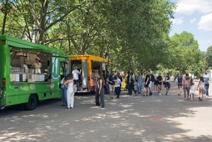 Ein grüner und ein gelber Foodtruck steht unter großen Platanen, davor stehen Menschen Schlange und laufen vorbei