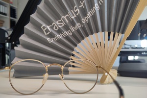 Im Vordergrund liegt eine Brille, im Hintergrund ein aufgefächerter grauer Papierfächer mit Aufschrift "Erasmus+: Enriching lives, opening minds."