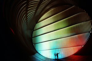 Ein Mensch steht in einem Windkanal, der in Licht in allen Farben des Regenbogens getaucht ist.