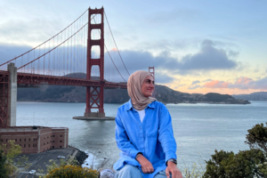 Eine junge Frau mit blauer Bluse und hellem Kopftuch sitzt im Vordergrund, hinter ihr eine große rote Brücke über ein großes Gewässer. Der Horizont ist von der Abend- oder Morgendämmerung rötlich gefärbt.