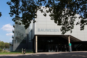 Foto eines grauen Gebäudes, das Wort "Bauwesen" steht in Großbuchstaben darauf. Im Vordergrund steht ein Baum, an der Seite des Gebäudes ist ein Treppenhaus zu sehen.
