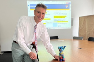 Ein Mann in Hemd und Krawatte hält die Hand an eine auf einem langen Konferenztisch stehende Bärenfigur, im Hintergrund ist eine Präsentation an die Wand gebeamt. 