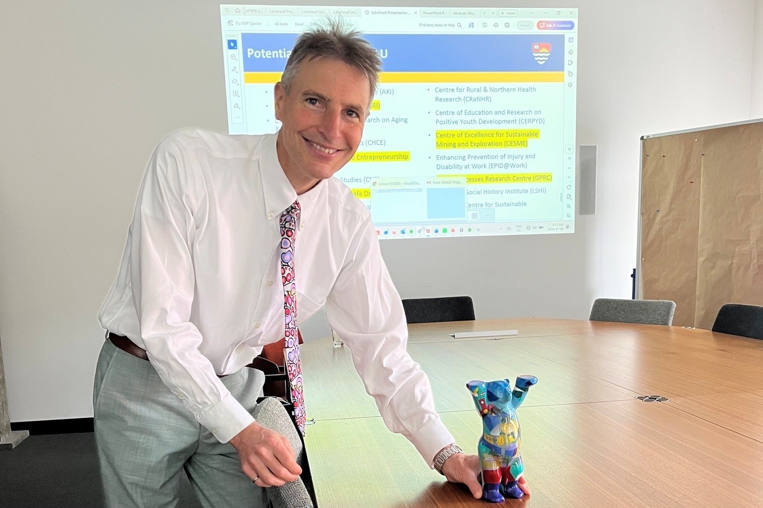 Ein Mann in Hemd und Krawatte hält die Hand an eine auf einem langen Konferenztisch stehende Bärenfigur, im Hintergrund ist eine Präsentation an die Wand gebeamt. 