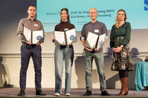 Zwei Männer und eine Frau mit Urkunden in den Händen sowie eine weitere Person stehen nebeneinander auf einer Bühne