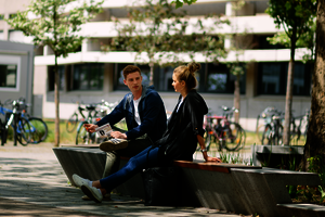 Zwei Personen sitzen auf einer Bank auf dem Campus. Im Hintergrund Bäume und Fahrräder.