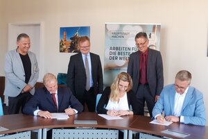 Drei Personen sitzen an einem Tisch und unterschreiben jeweils ein Dokument, dahinter stehen drei weitere Personen 