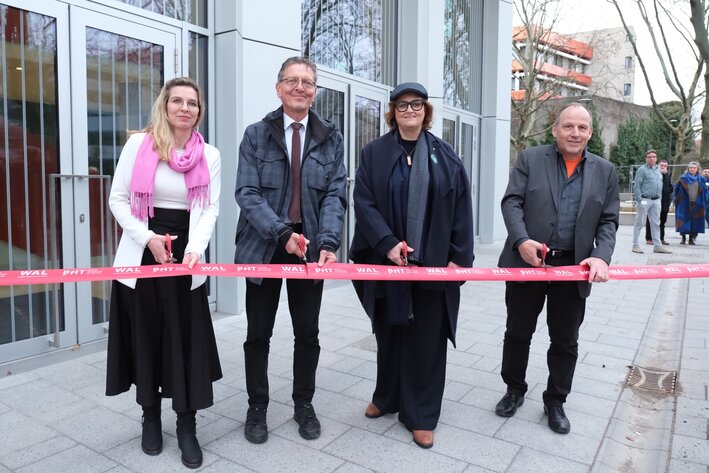 Vier Personen stehen vor einem Gebäude und halten Scheren bereit, um ein rotes Band durchzuschneiden
