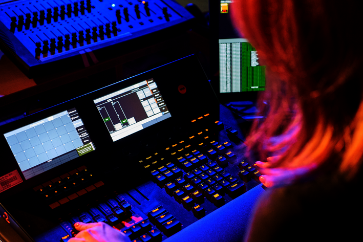 Eine Person steht an einem Technikpult mit Displays und hantiert an den Reglern.