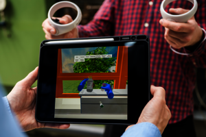 Ein Student steuert per Controller blaue Hände in einem virtuellen Labor. Der Vorgang im virtuellen Raum wird durch ein Tablet beobachtet.