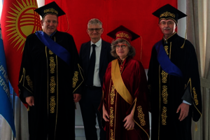 Vier Professor*innen posieren vor einer roten Flagge mit Sonnensymbol. Drei von ihnen tragen Roben und Doktorhüte.