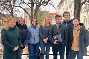 Sieben Personen stehen nebeneinander, im Hintergrund der winterliche Campus mit Schnee und kahlen Bäumen