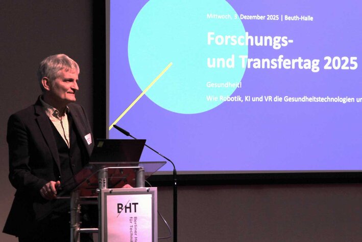 Mann steht am Rednerpult auf der Bühne, hinter ihm ist die Projektion einer Präsentation zu sehen