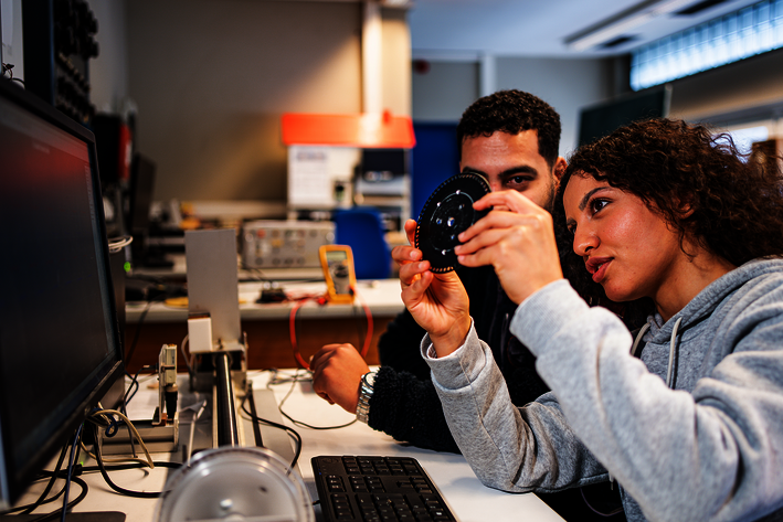 Zwei Studierende betrachten ein runde Bauteil. Im Hintergrund sind Arbeitsplätze und technische Geräte zu sehen.