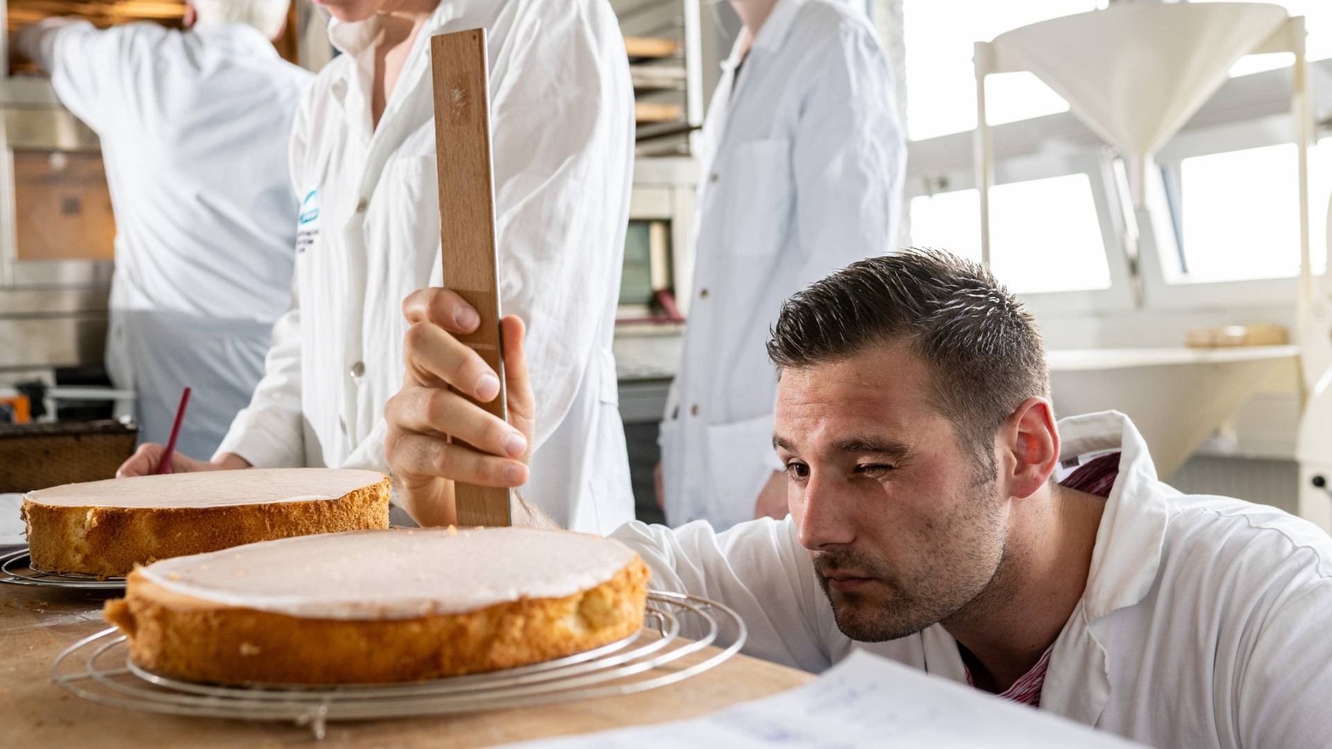 Ein Mann in weißem Kittel misst mit einem Holzlineal die Höhe eines Tortenbodens