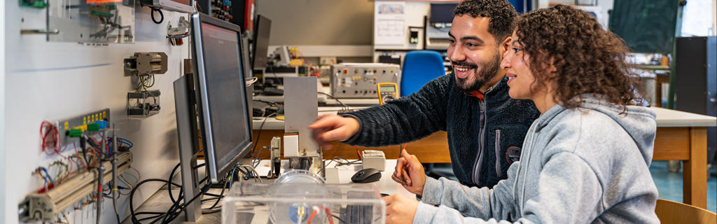Ein junger Mann und eine junge Frau schauen auf einen Bildschirm. Auf dem Tisch vor dem Bildschirm sowie an den Wänden und auf einem weieren Tisch im Raum sind Kabel, Prüfgeräte und weitere Ausstattungsgegenstände des elektrotechnischen Labors zu sehen.