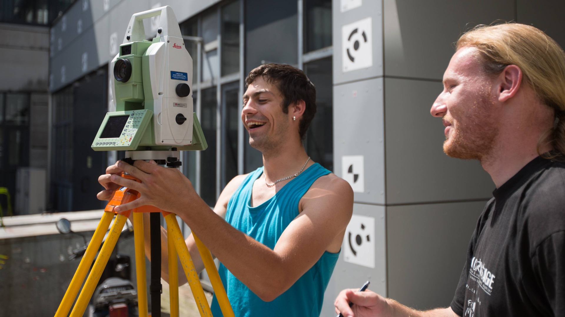 Zwei Männer richten auf dem Campus lachend Geräte zur Vermessung ein