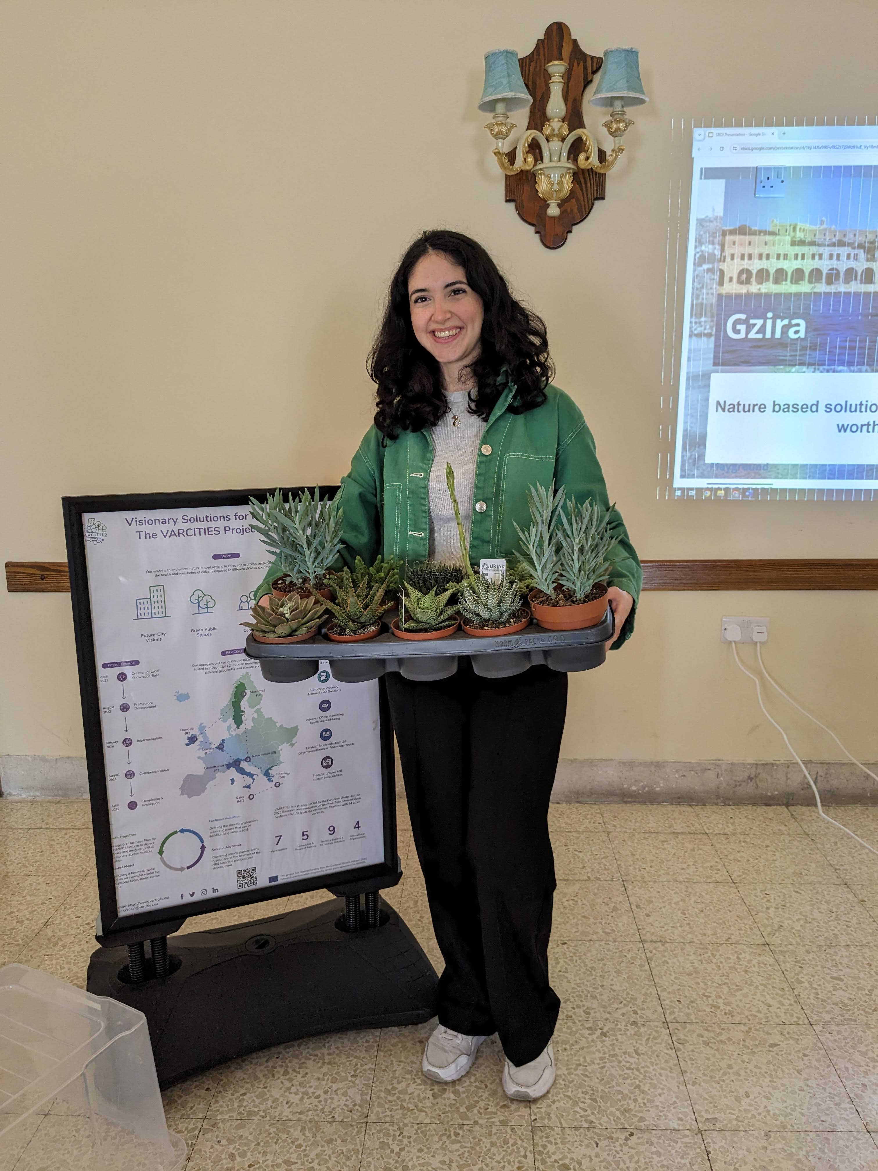 Eine Frau hält eine Palette mit Sukkulenten-Töpfen, sie steht vor einer Info-Tafel, im Hintergrund ist die Projektion einer Präsentation zu sehen