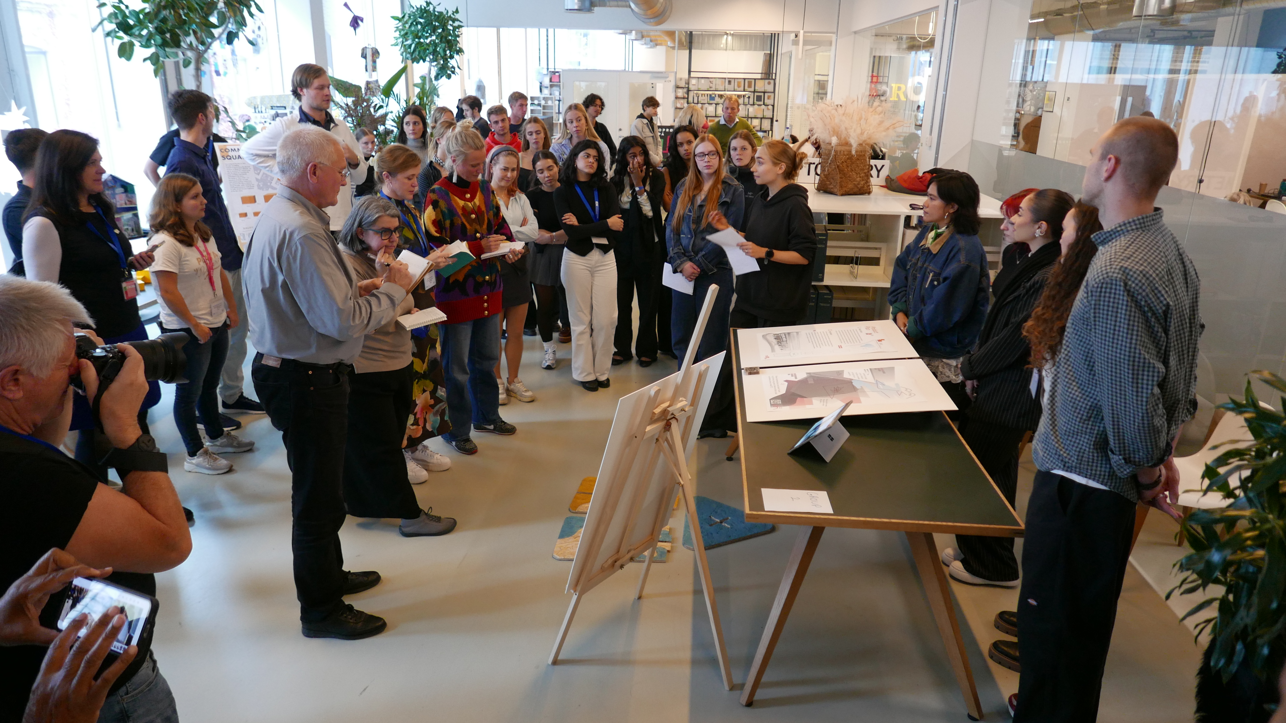 Five students behind a table seem to be presenting some sketches or visualizations ot a larger group of people.