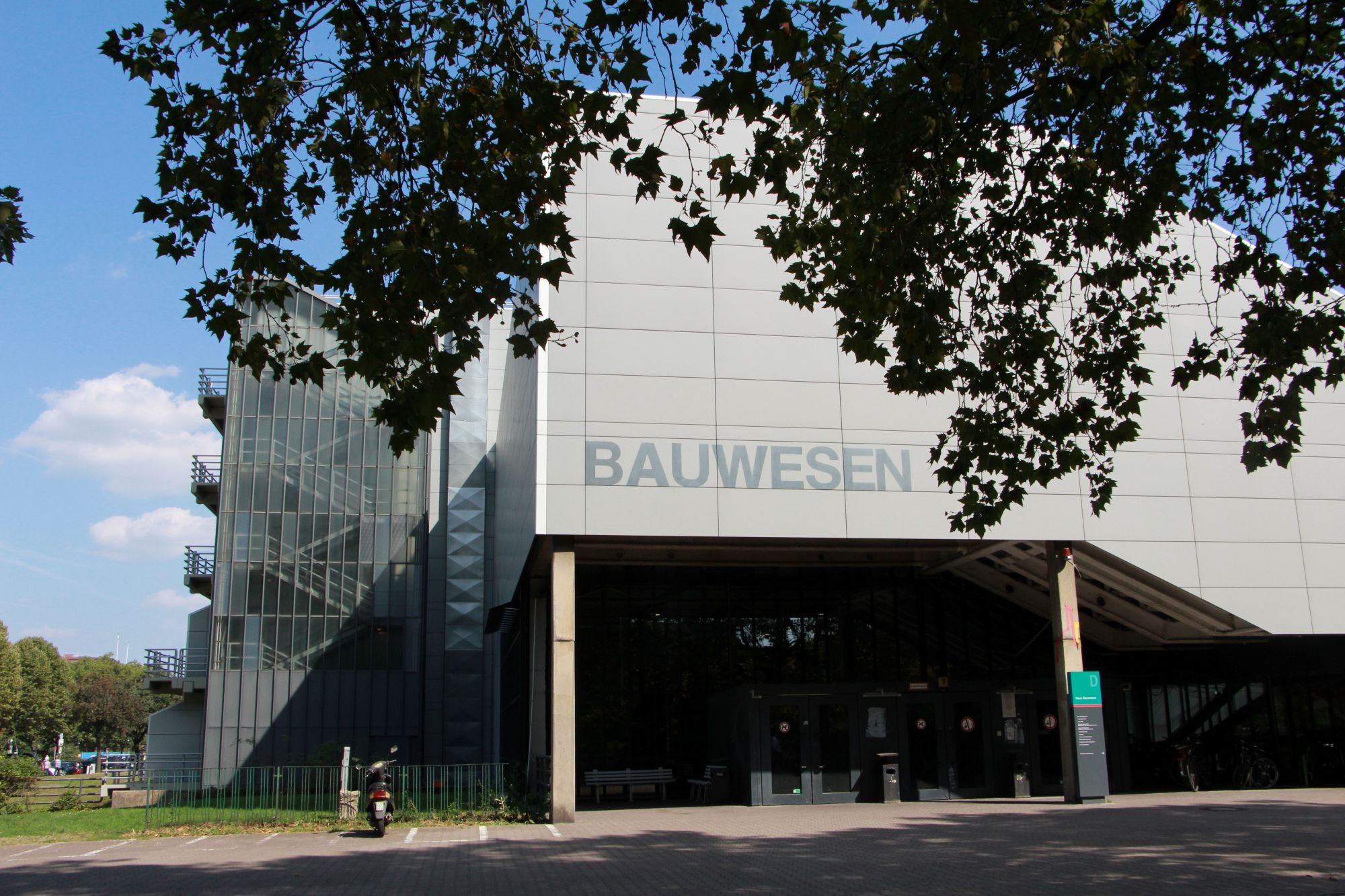 Foto eines grauen Gebäudes, das Wort "Bauwesen" steht in Großbuchstaben darauf. Im Vordergrund steht ein Baum, an der Seite des Gebäudes ist ein Treppenhaus zu sehen.