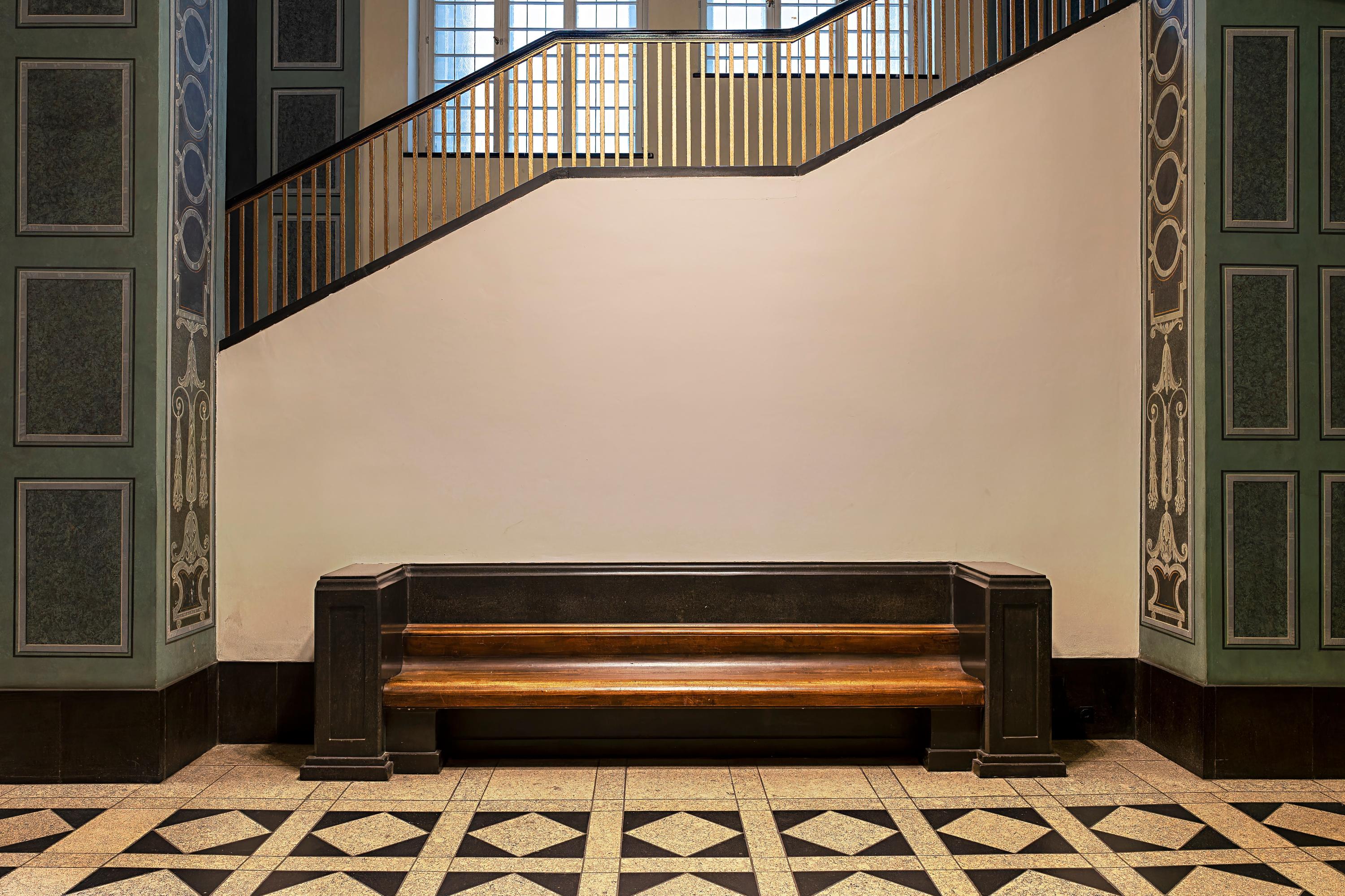 Eine hölzerne Bank steht vor einem Treppenaufgang zwischen zwei grünen, ornamentierten Säulen. Der Fußboden ist gefließt, im Hintergrund sind zwei große Sprossenfenster im Anschnitt zu sehen.
