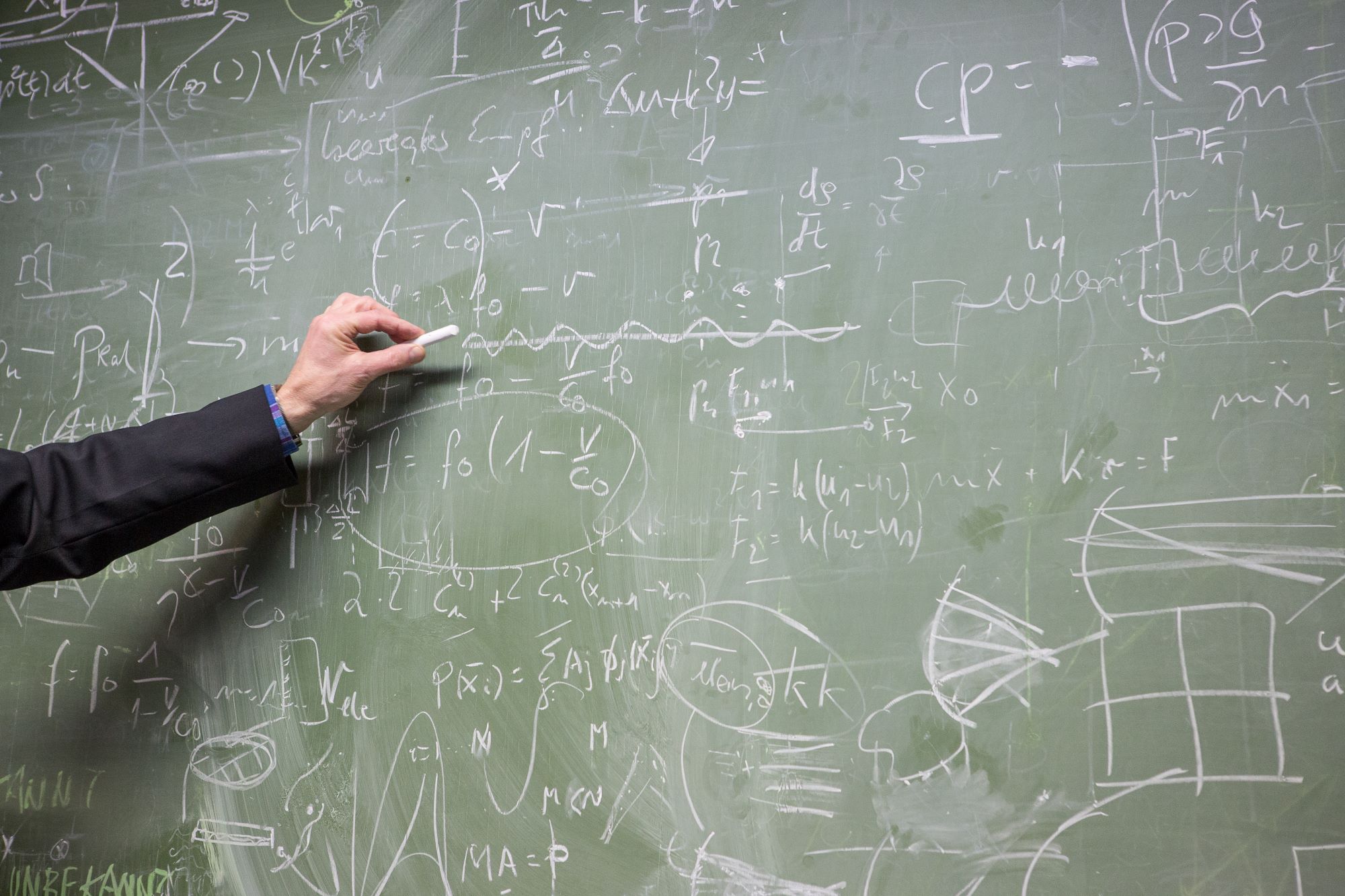 Foto einer mit Formeln vollgeschriebenen Tafel. Von links ragt der Arm eines Menschen hinein, der ein Stück Kreide in der Hand hält.