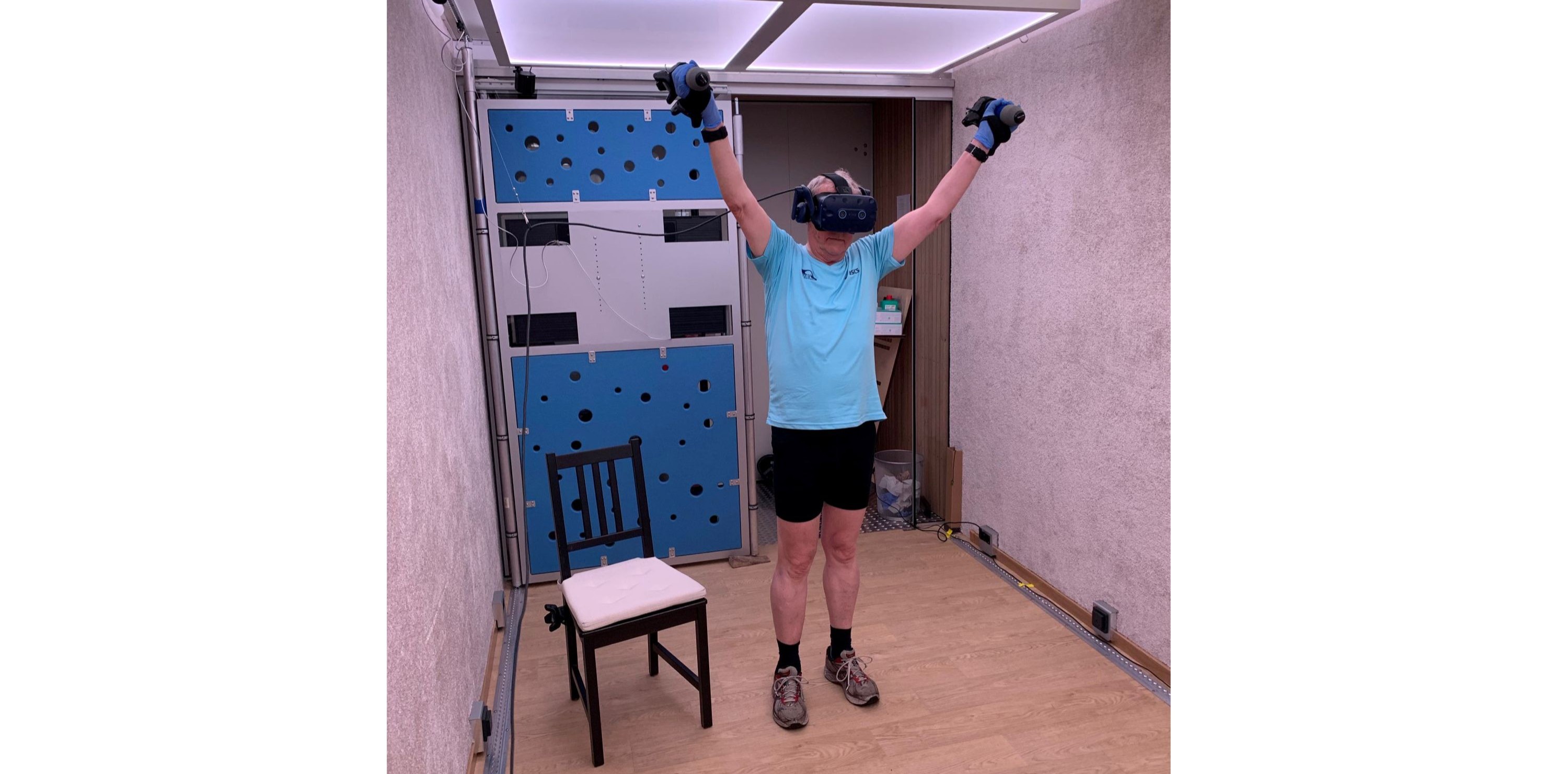 Eine ältere Person in Sportkleidung steht in einem kleinen Raum und trägt eine VR-Brille. Sie streckt die Arme nach oben und hält in jeder Hand einen Controller. Links neben ihr steht ein Stuhl, hinter ihr eine blaue Wand.