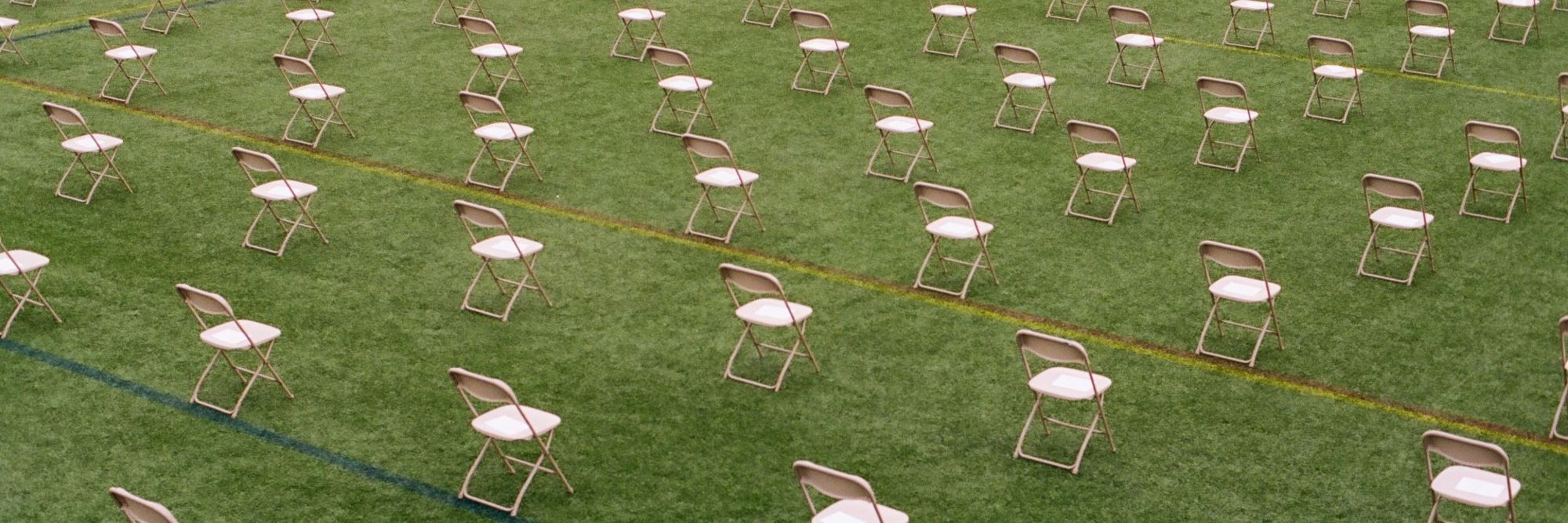 Weiße Stühle stehen mit viel Abstand zueinander in Reihen auf einer Wiese.