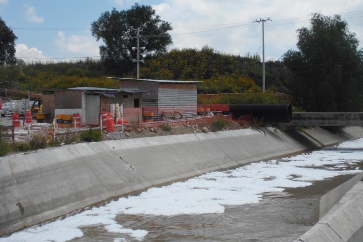 Ein Kanal mit dunklem Wasser, auf dem sich stellenweise Schaum befindet. Daneben eine Wellblechhütte, im Hintergrund Hecken und Bäume.
