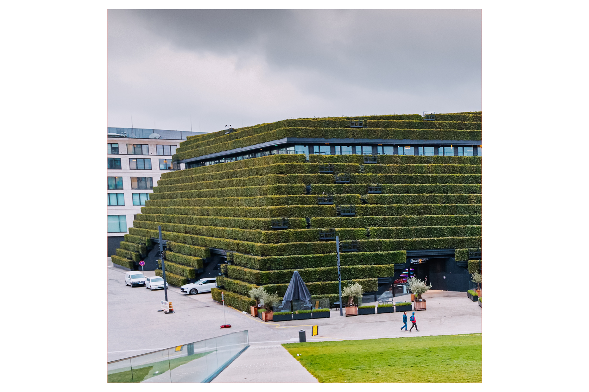 Blick aus der Vogelperspektive auf ein Gebäude mit schräger und in Stufen umlaufender Fassade, die komplett begrünt ist. Davor eine Grünfläche und Menschen, rechts reicht der Blick auf eine rechst um die Kurve laufende Straße