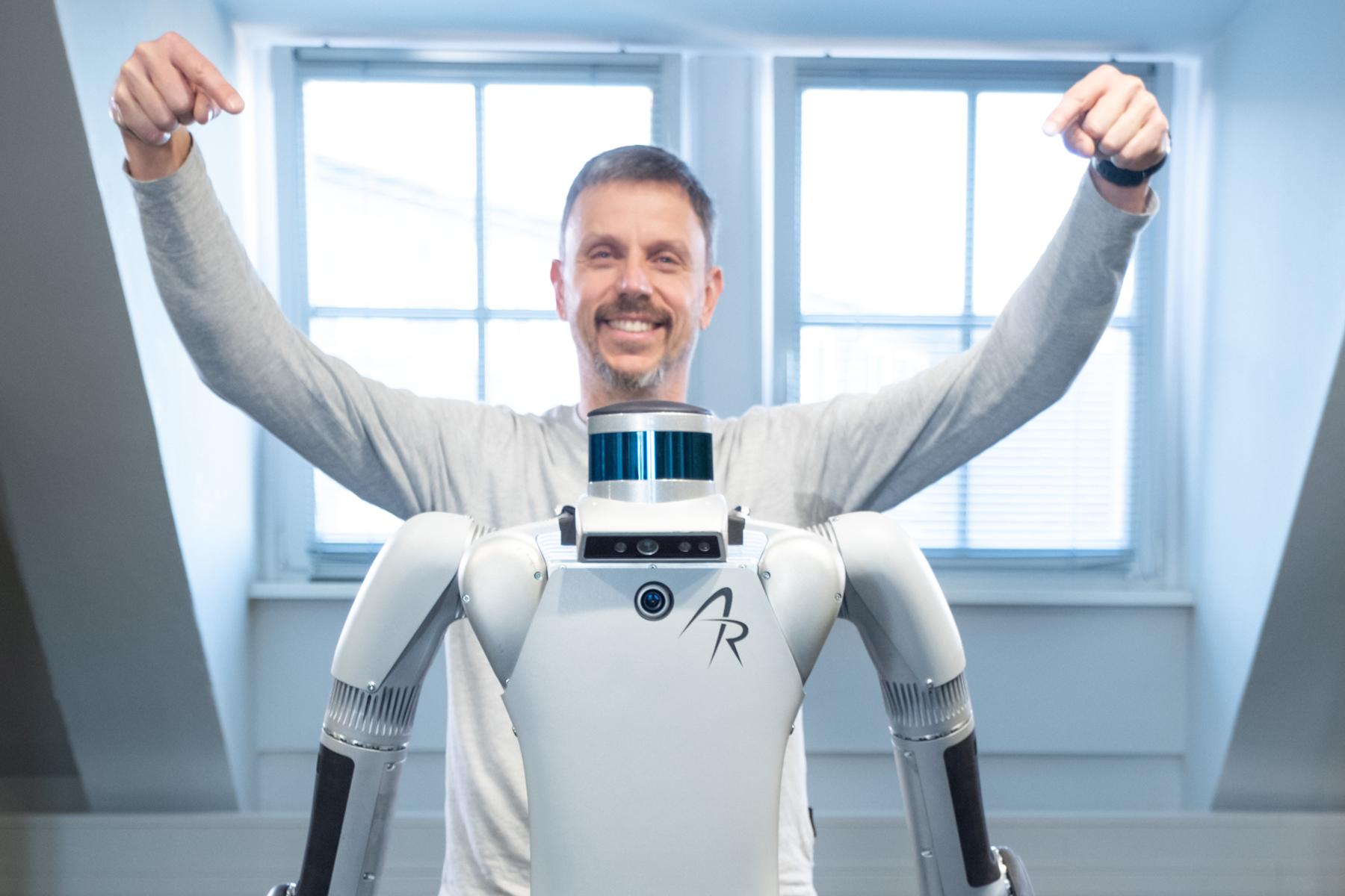 Ein Mann steht hinter einem Roboter und zeigt mit erhobenen Armen auf ihn