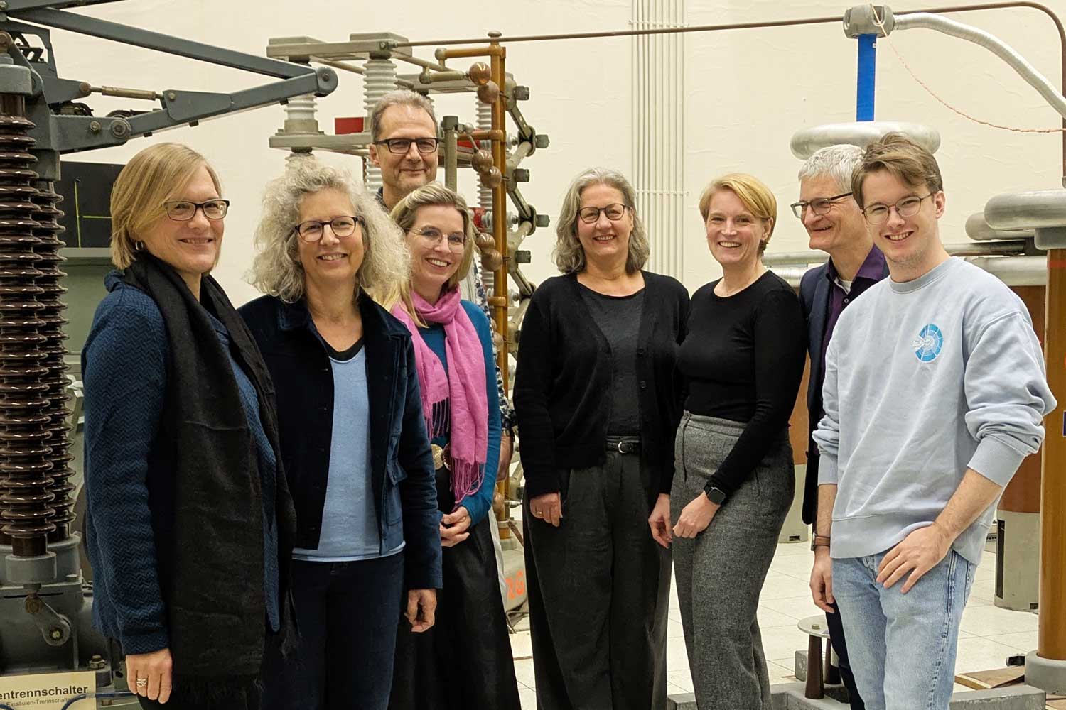 Acht Männer und Frauen stehen als Gruppe vor elektronischen Apparaturen