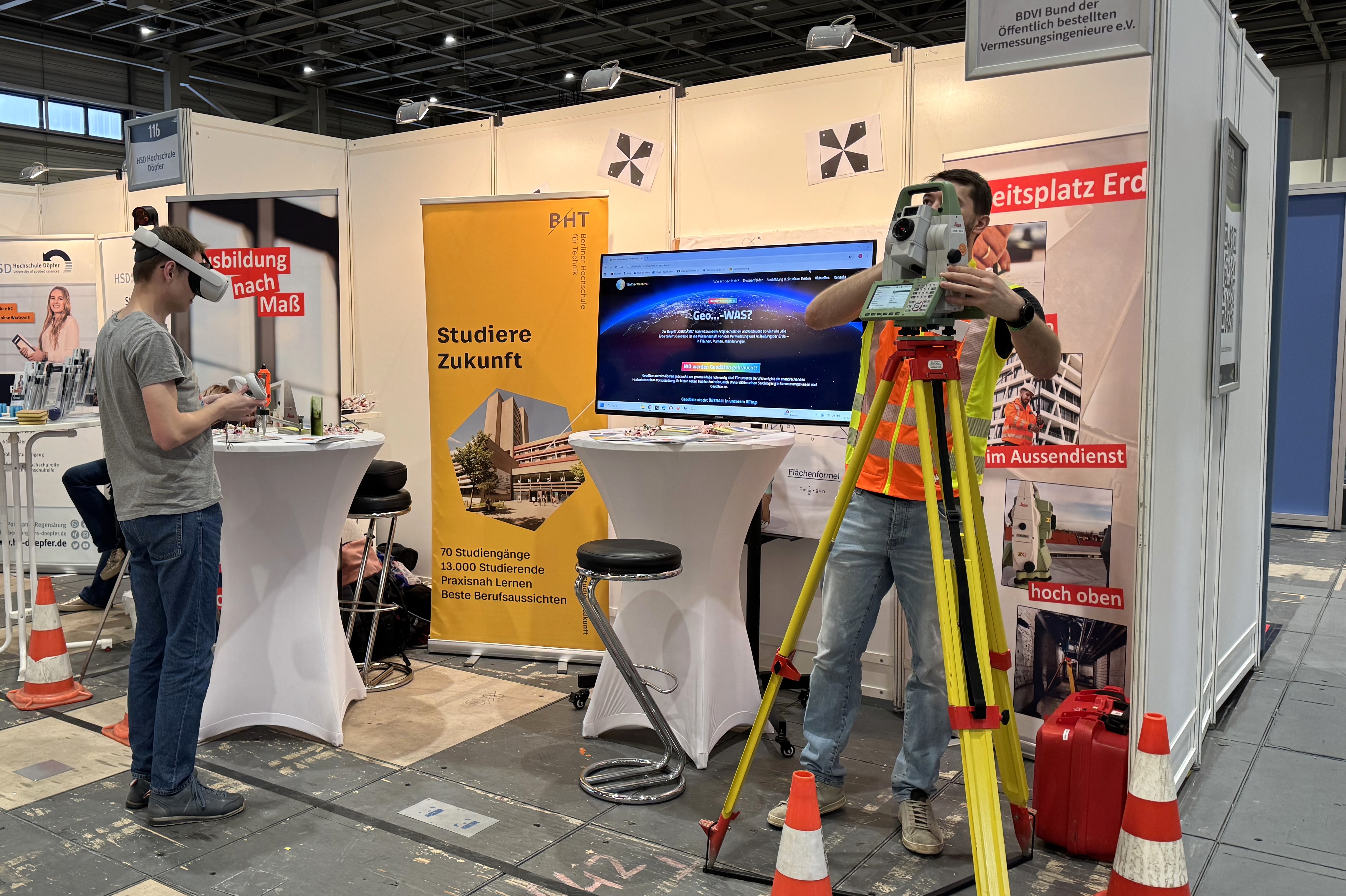 Vor einem Messestand blickt ein Student durch ein Vermessungsgerät, ein anderer trägt eine VR-Brille
