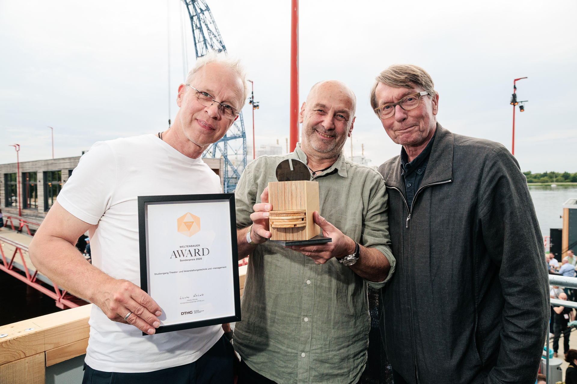 Drei Männer posieren auf einem Hafengelände mit einer gerahmten Urkunde und einem Award