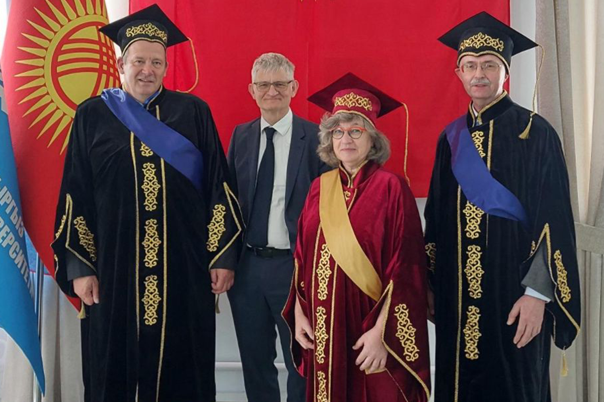 Vier Professor*innen posieren vor einer roten Flagge mit Sonnensymbol. Drei von ihnen tragen Roben und Doktorhüte.