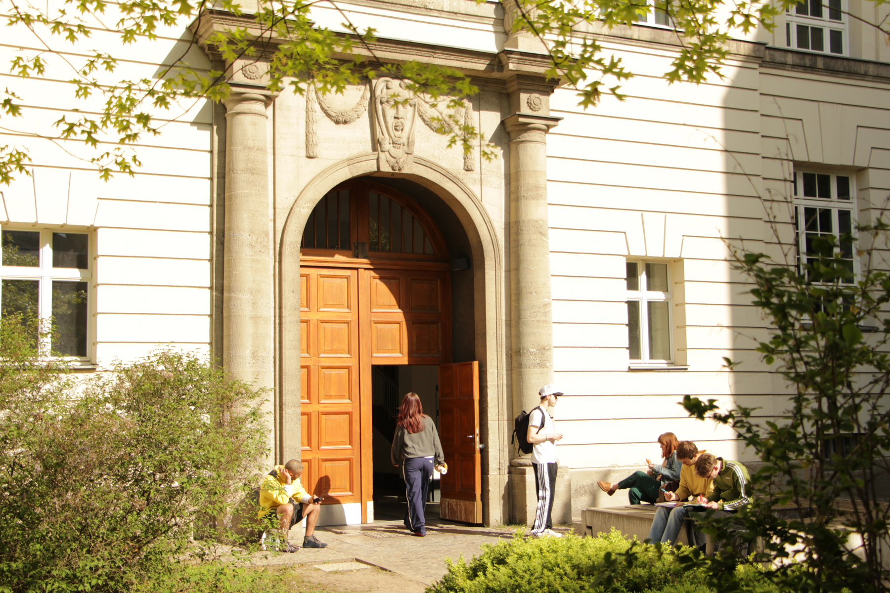 Eingangsbereich des Hauses Beuth der BHT umgeben von grünen Bäumen und Büschen, davor Studierende in der Sonne