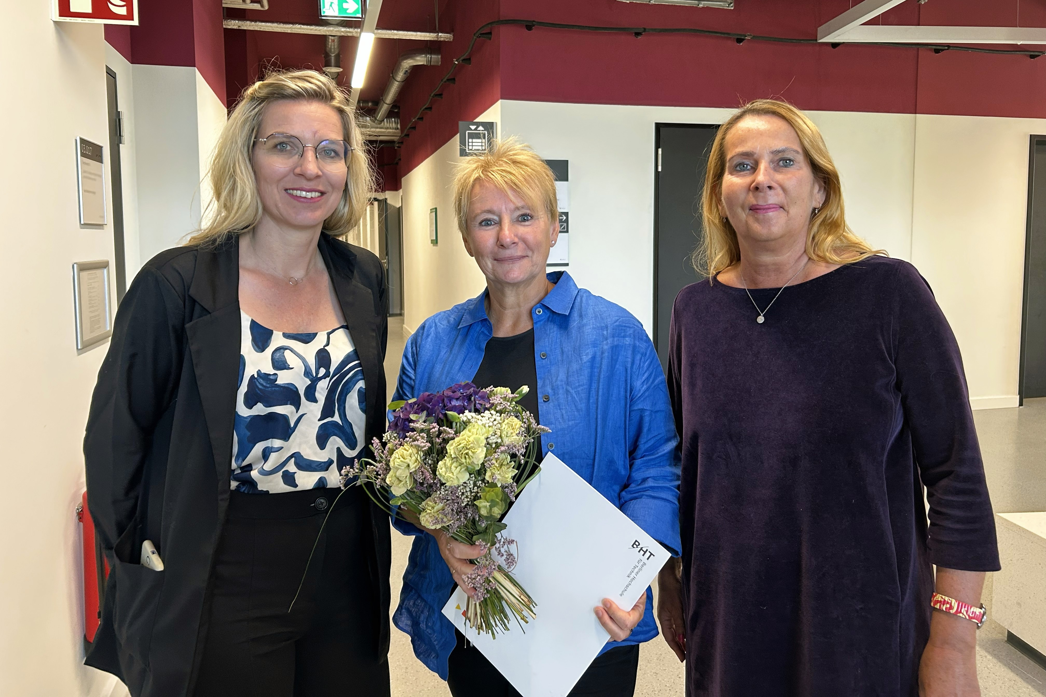 Drei Frauen blicken in die Kamera, Kristin Grzedzinski in der Mitte hält einen Blumenstrauß und eine Urkunde in den Händen. 