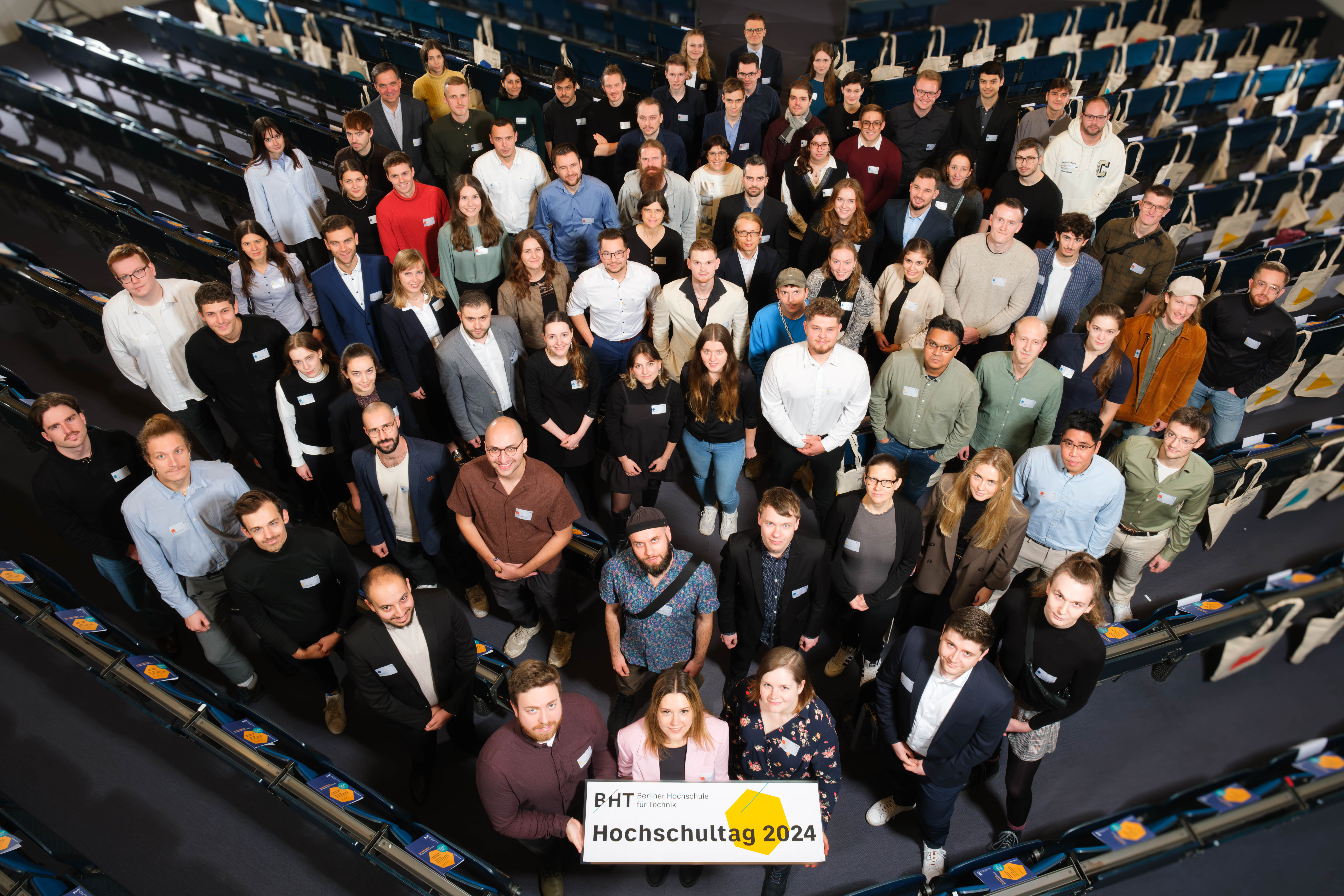 Eine große Gruppe an Personen steht in Reihen eines Hörsaals. Die Personen vorne halten ein Schild mit den Worten Hochschultag 2024.