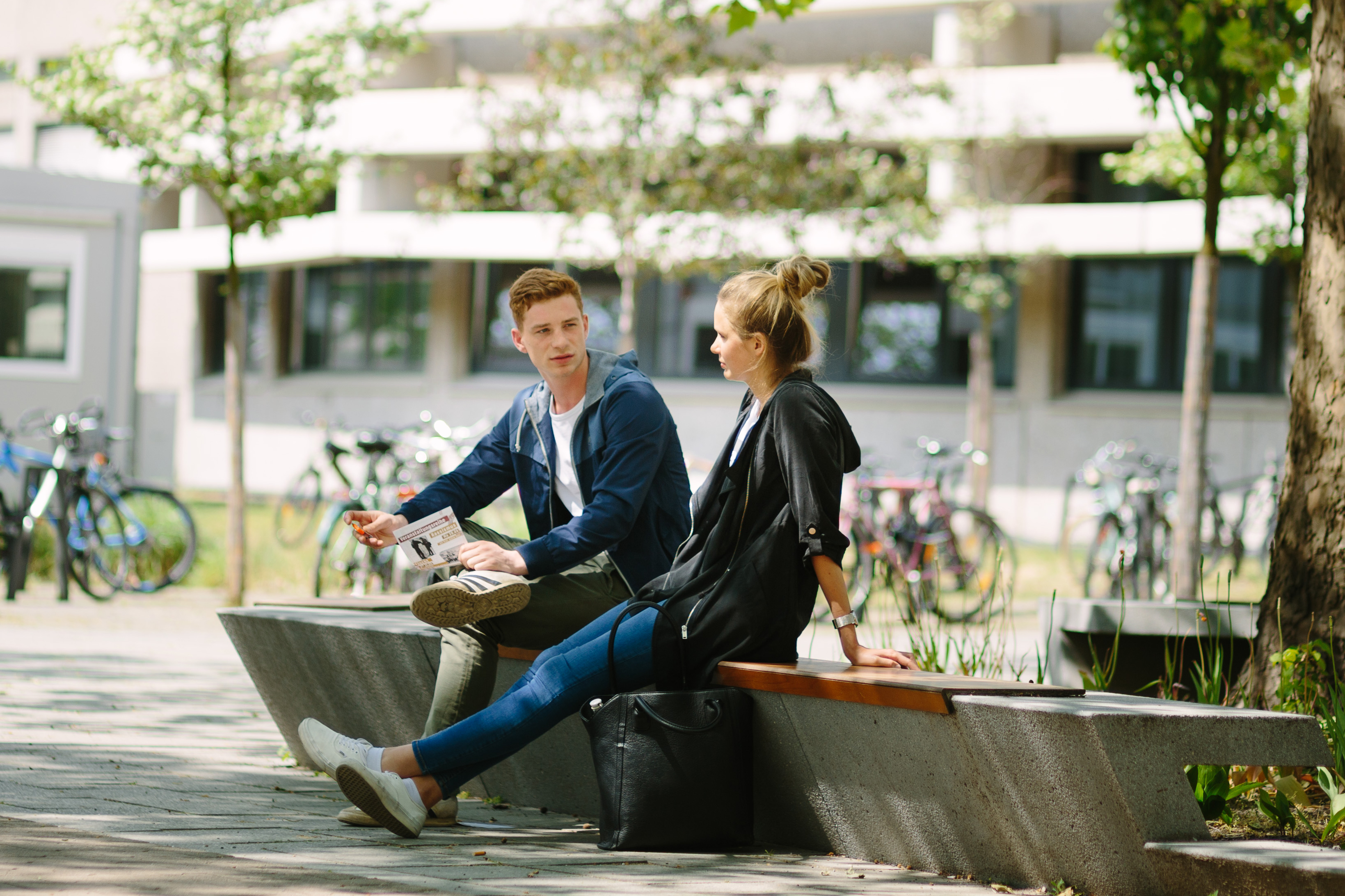 Zwei Personen sitzen auf einer Bank auf dem Campus. Im Hintergrund Bäume und Fahrräder.