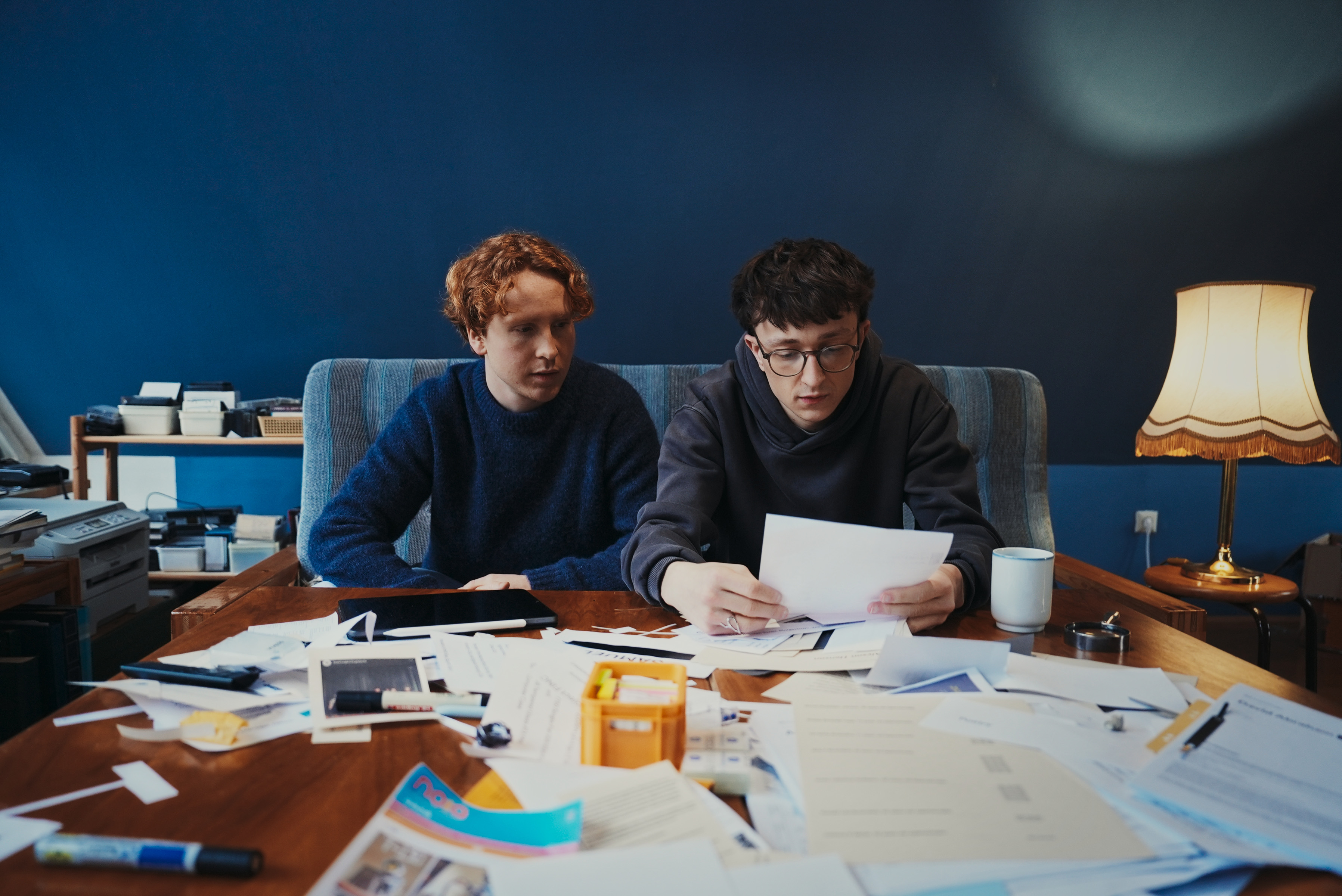 Zwei Männer sitzen an einem Tisch mit vielen Unterlagen und schauen auf ein Dokument