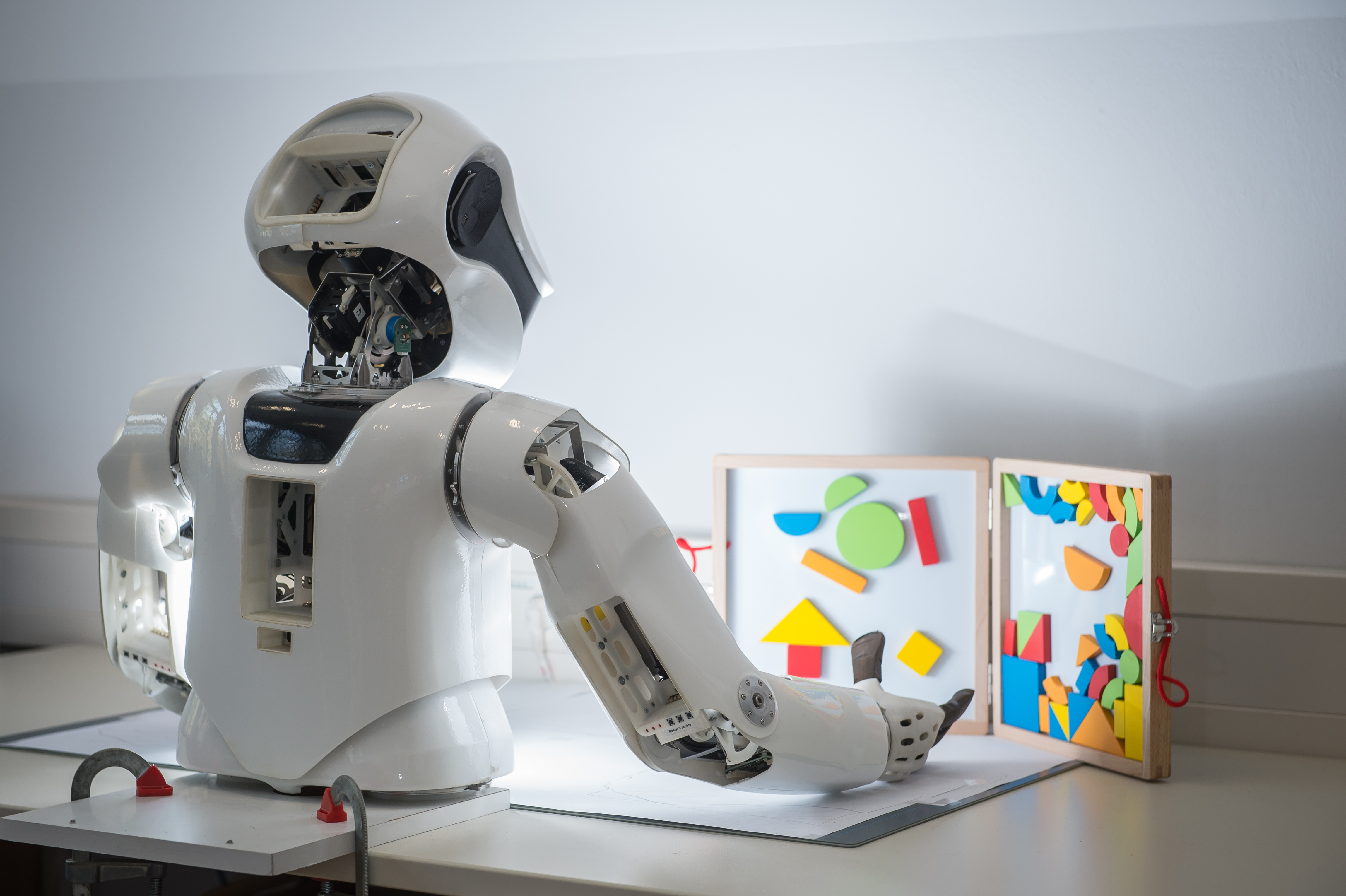 Rückansicht eines humanoiden Roboter-Oberkörpers, der auf einem Tisch steht und mit einer Hand nach einer Form auf einer Magnettafel greift