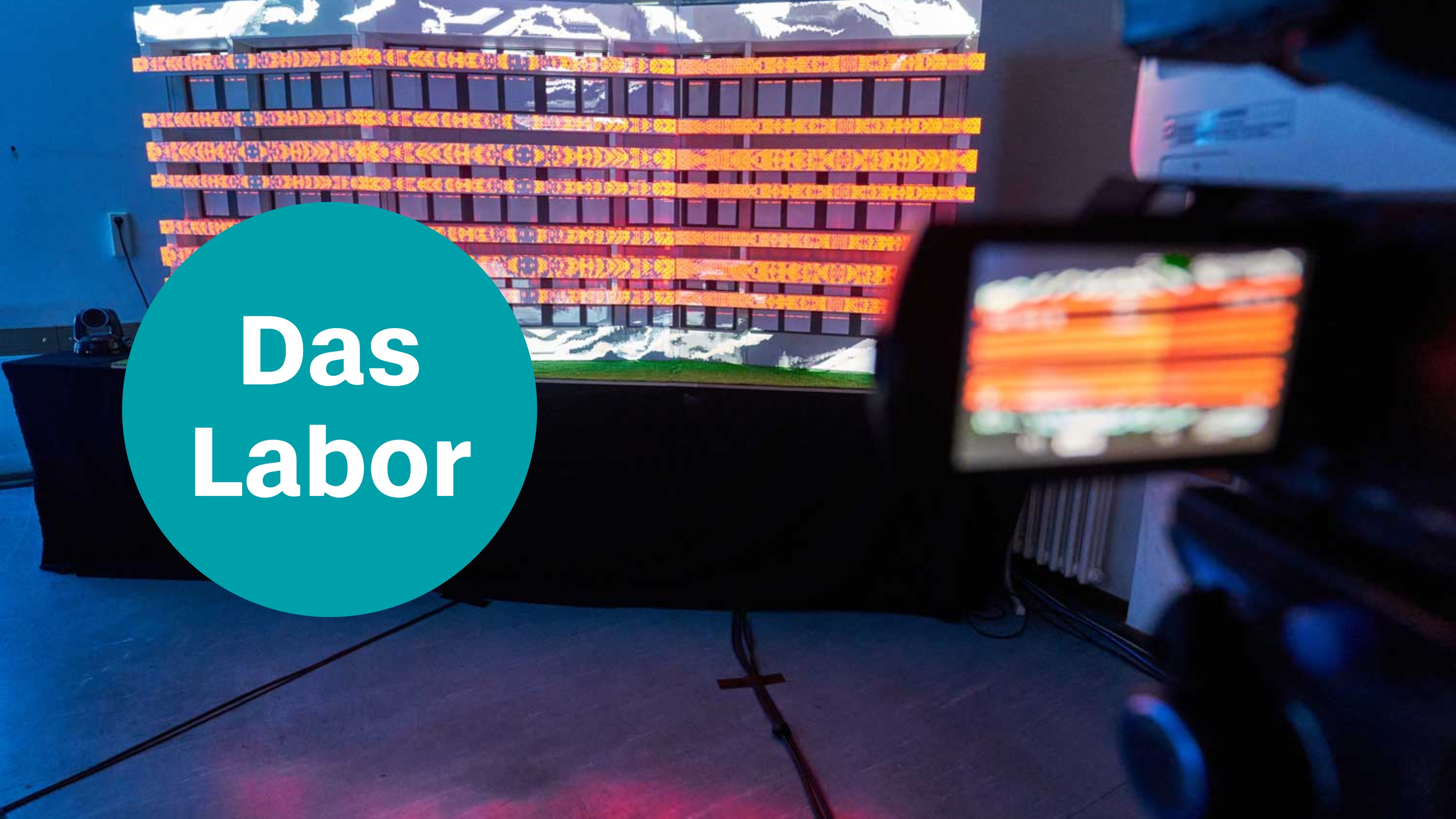 Ein illuminiertes Bühnenbild-Modell, das von einer Kamera mit Bildschirm gefilmt wird, Text: Das Labor