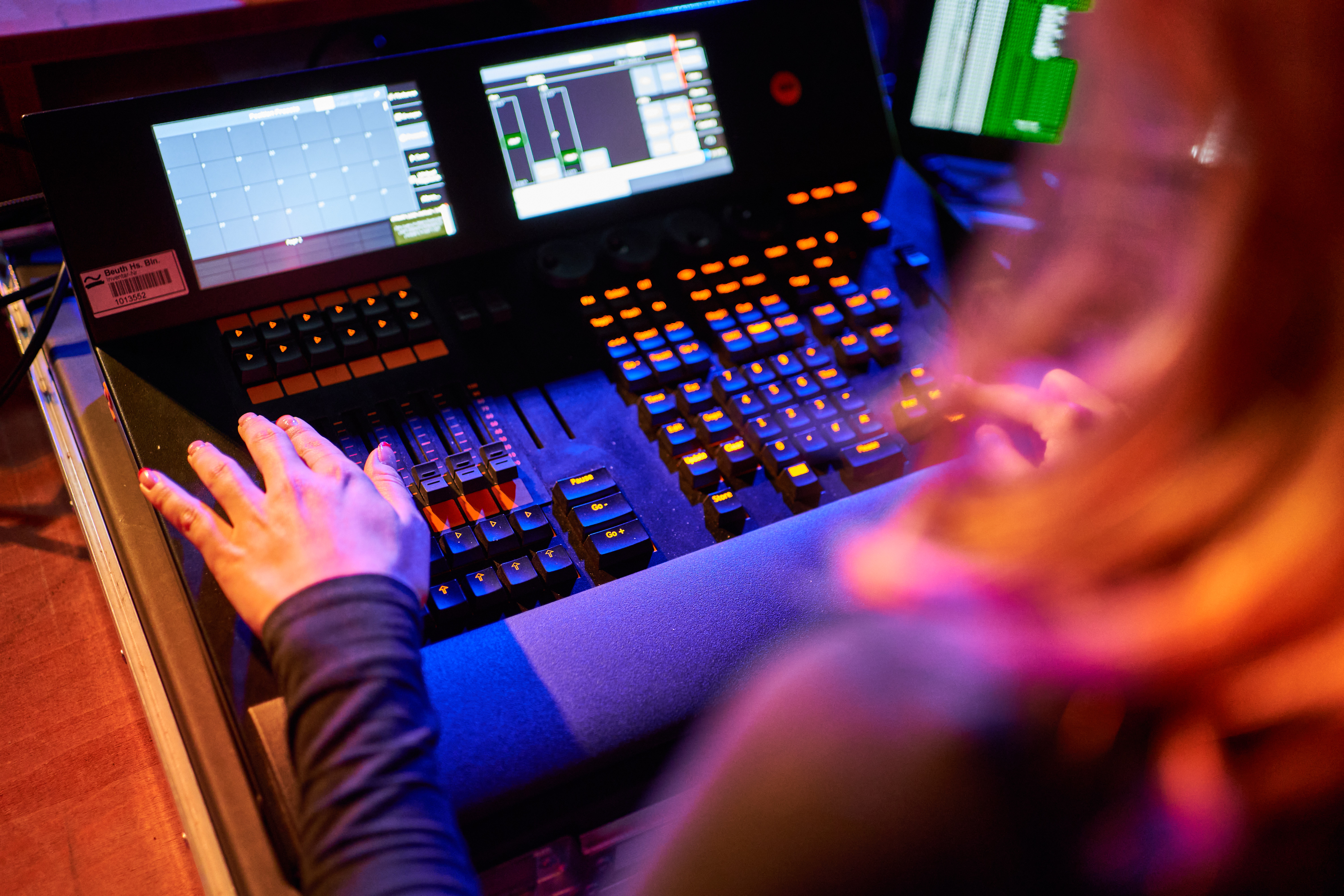 Blick über die Schulter einer Person, die ein Steuerungsboard für Bühnentechnik mit leuchtenden Knöpfen und Bildschirmen bedient