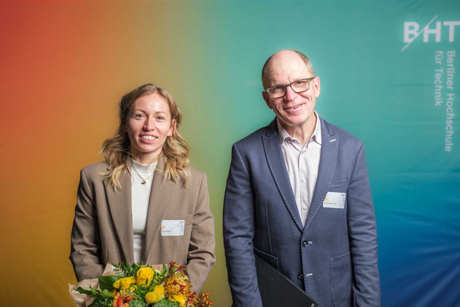 Frau mit Blazer und Mann in Anzug stehen nebeneinander, die Frau hält einen Blumenstrauß in der Hand
